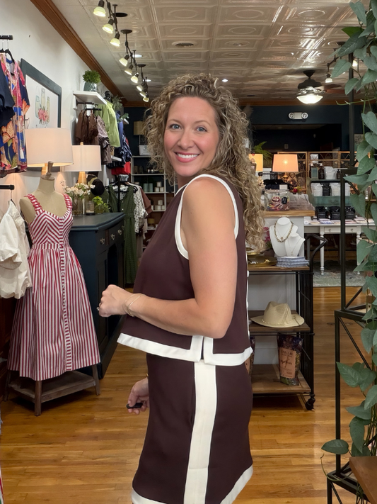 A woman with curly hair wearing a sleeveless brown dress with white contrast trim stands inside a retail boutique, smiling and looking over her shoulder. The store has wood floors, clothing racks, mannequins, and decorative plants.