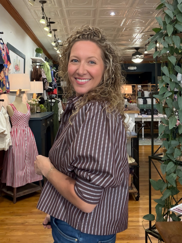 A smiling woman with curly hair wearing a brown and white vertically striped short-sleeve blouse and blue jeans, standing inside a boutique store.