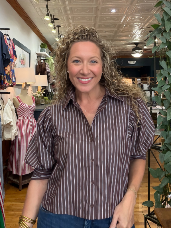 A smiling woman with curly blonde hair wearing a brown and white vertically striped short-sleeve button-up shirt, standing inside a boutique store.