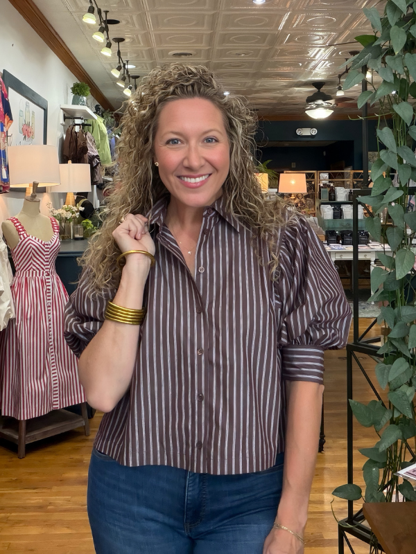 A smiling woman with curly brown hair stands in a boutique wearing a brown and white vertically striped short-sleeve button-up shirt with puffed sleeves and dark blue jeans.