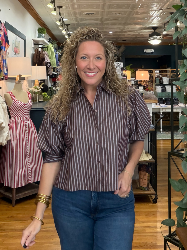A smiling woman with curly hair stands in a boutique wearing a brown and white vertically striped short-sleeve button-up shirt with blue jeans.