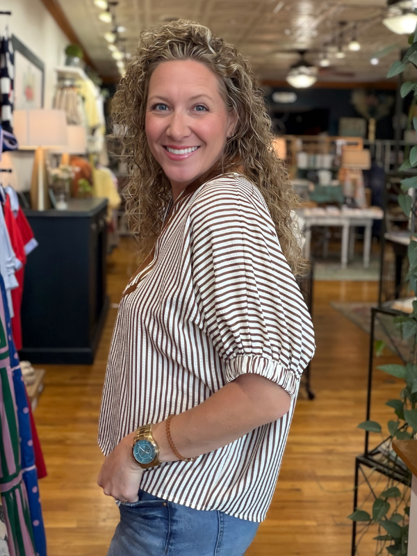 Woman in a striped shirt standing in a store with clothing and decor items around.