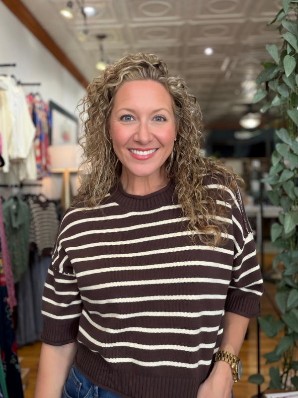 A smiling woman with curly blonde hair wearing a brown and cream horizontally striped short-sleeve knit sweater, standing inside a clothing boutique.