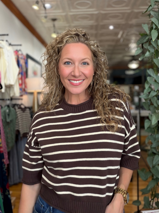 A smiling woman with curly blonde hair wearing a brown and cream horizontally striped short-sleeve knit sweater, standing inside a clothing boutique.