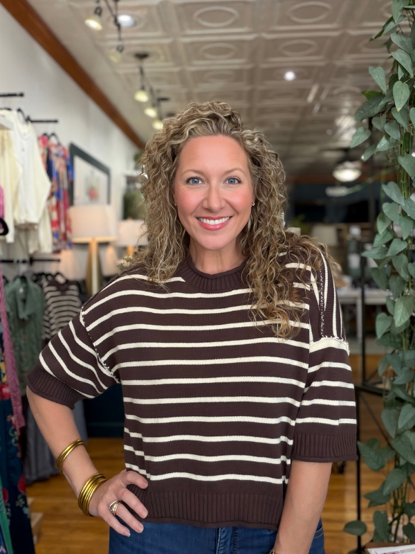 A smiling woman with curly blonde hair wearing a brown and cream horizontally striped knit sweater, standing in a clothing boutique.