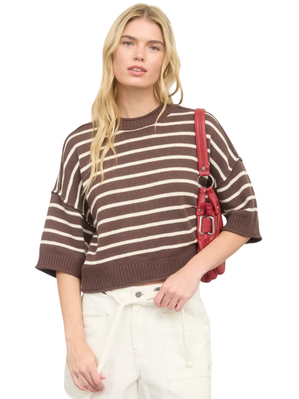 A blonde model wearing a brown and cream striped knit sweater, white pants, and carrying a red shoulder bag against a plain white background.