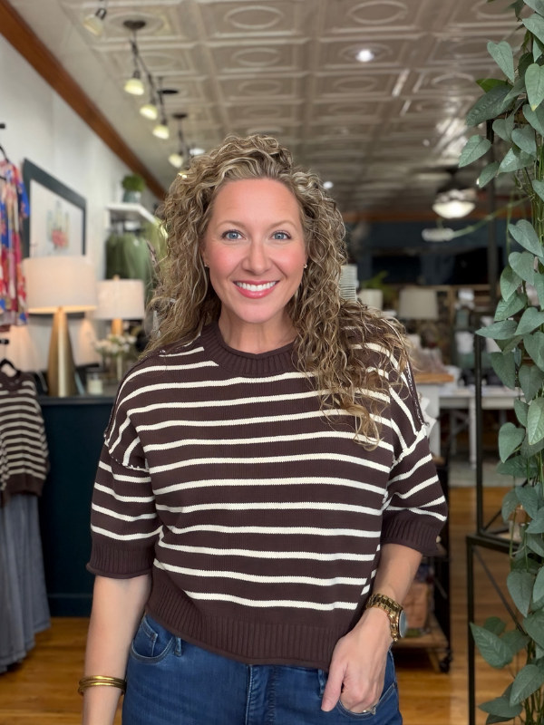 A smiling woman with curly blonde hair wearing a brown and cream horizontally striped knit sweater and blue jeans, standing inside a boutique store.