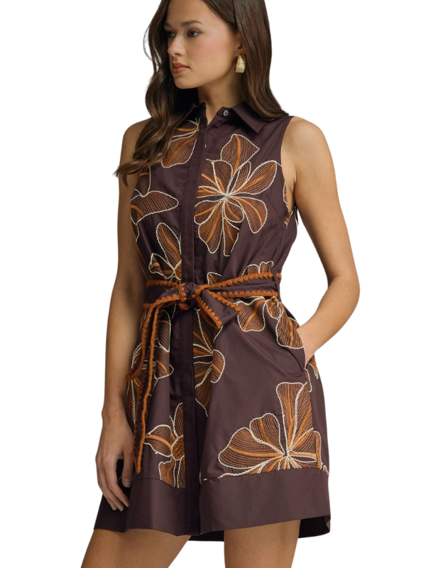 A woman modeling a sleeveless brown mini dress with a brown and beige floral print, featuring a button-up front, collar, and a braided brown tie belt at the waist.
