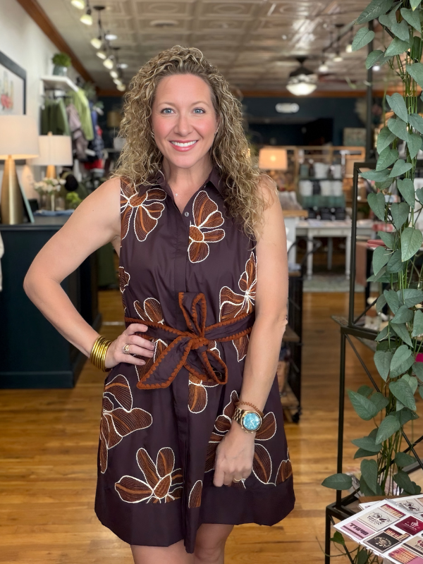 A smiling woman with curly blonde hair stands in a boutique wearing a sleeveless dark brown dress with a large brown and white floral pattern and a matching fabric tie belt at the waist.