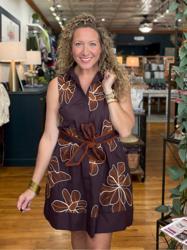 A woman with curly blonde hair wearing a sleeveless dark brown mini dress with a large brown and white floral pattern and a matching textured brown belt, standing in a retail store.