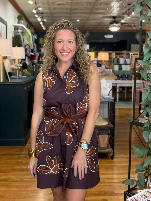 A smiling woman with curly blonde hair stands in a boutique wearing a sleeveless dark plum mini dress with a brown and beige floral pattern and a matching fabric tie belt.