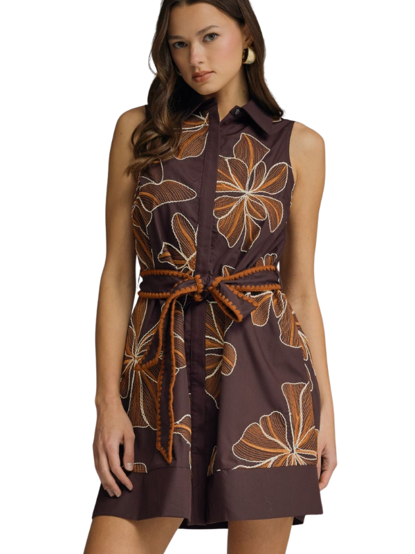 A woman modeling a sleeveless brown shirt dress with a large tan and white floral print, featuring a matching braided tie belt at the waist.