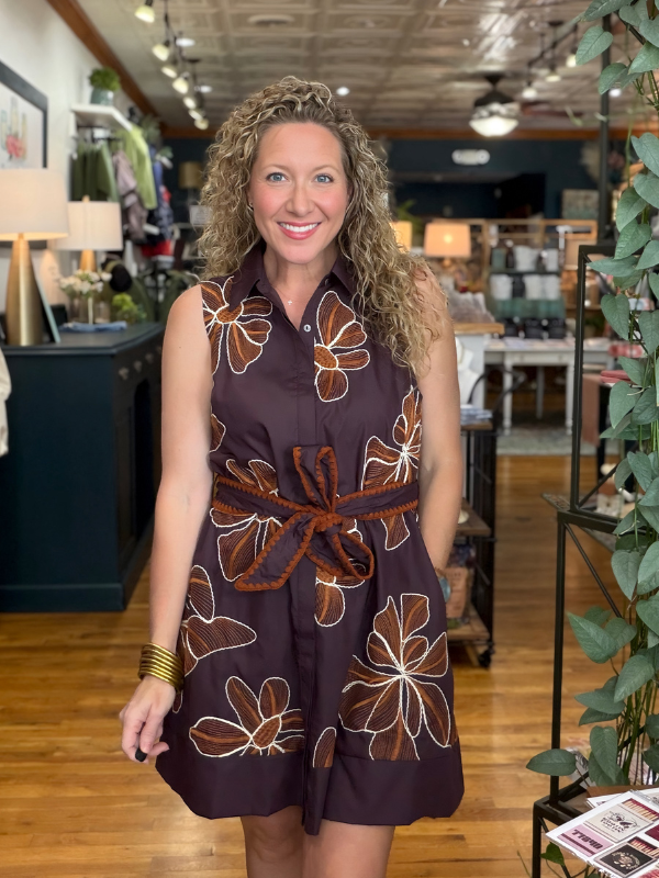 A woman with curly blonde hair stands in a boutique wearing a sleeveless dark plum button-up dress with a brown and white floral pattern and a braided brown belt.