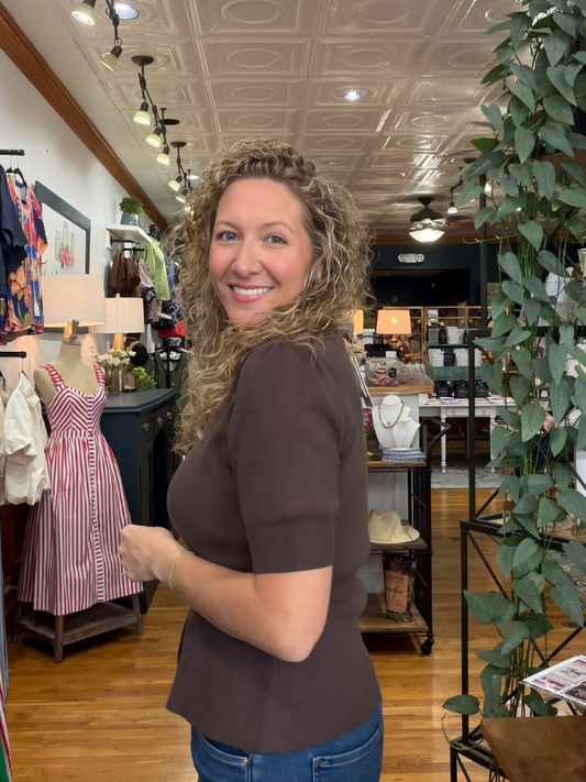 A smiling woman with curly hair wearing a brown short-sleeve top and blue jeans, standing inside a boutique store.