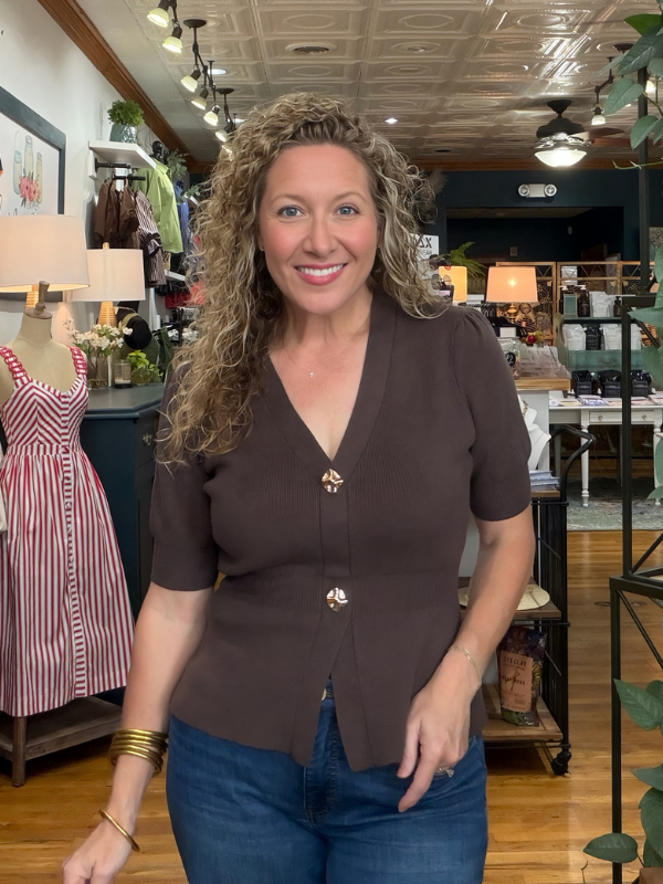 A smiling woman with curly hair stands in a boutique shop wearing a dark brown short-sleeve button-front top and blue jeans.