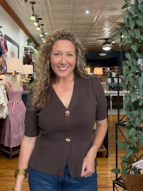 A smiling woman with curly blonde hair wearing a brown short-sleeve button-front top and blue jeans, standing inside a retail store with clothing racks, lamps, and a ceiling fan visible in the background.