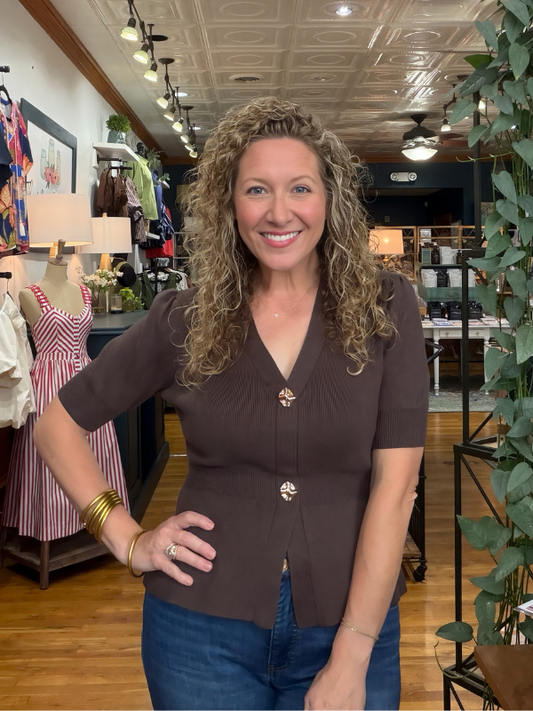 A smiling woman with curly blonde hair stands in a boutique wearing a brown short-sleeve knit top with decorative gold-tone buttons and blue jeans.