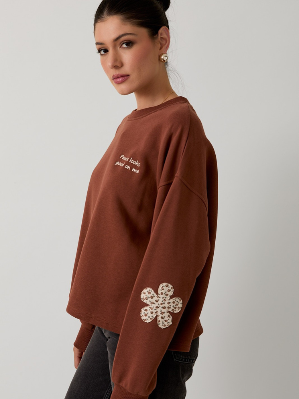 Woman wearing a brown sweatshirt with a white flower design on a plain background