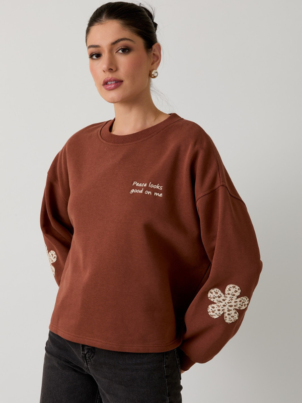 Woman wearing a brown sweatshirt with floral designs and text on a plain background