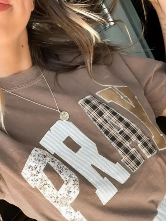 A close-up of a person wearing a brown graphic sweatshirt with large white and plaid lettering, along with a silver necklace.