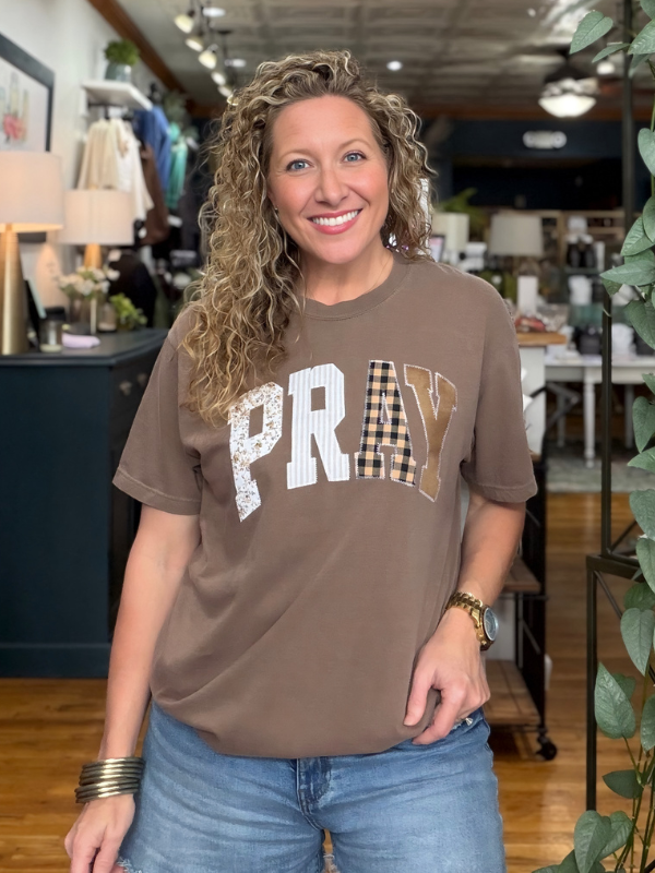 A smiling woman with curly blonde hair stands in a boutique wearing a brown graphic t-shirt with 'PRAY' written across the chest in white, textured, and gingham patterns, paired with blue jeans.