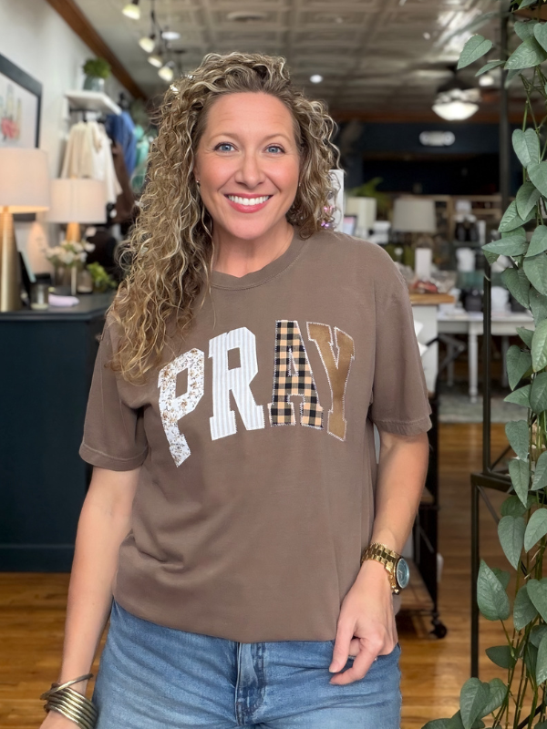 A smiling woman with curly blonde hair stands in a boutique wearing a brown graphic t-shirt with 'PRAY' written across the chest in white, textured, and gingham patterns, paired with blue jeans.