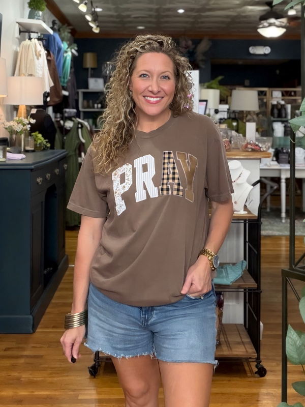 A smiling woman with curly blonde hair stands in a boutique wearing a brown graphic t-shirt with 'PRAY' written across the chest in white, textured, and gingham patterns, paired with blue jeans.