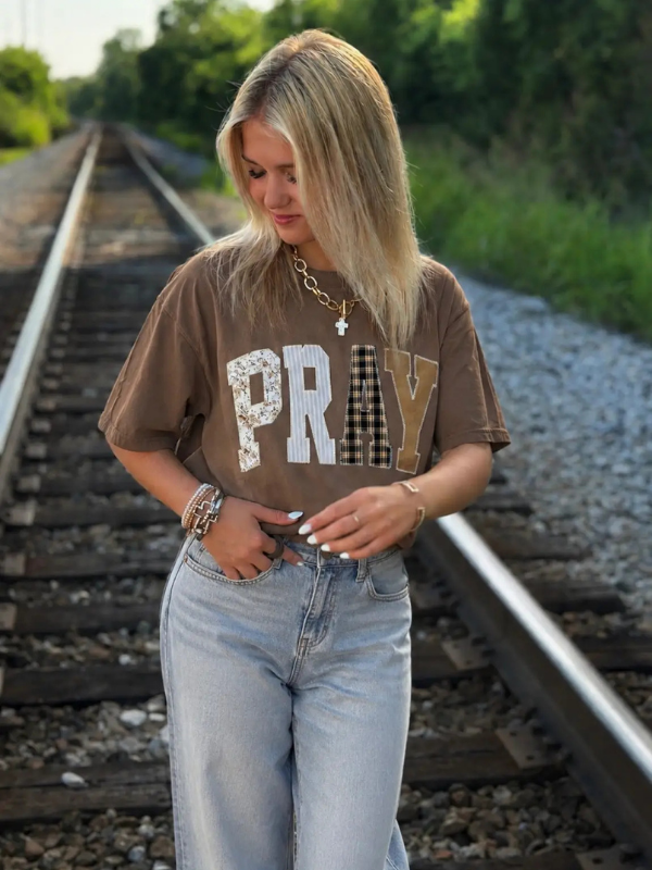 A woman stands on railroad tracks wearing a brown graphic t-shirt with the word 'PRAY' in textured lettering, paired with light-wash jeans.