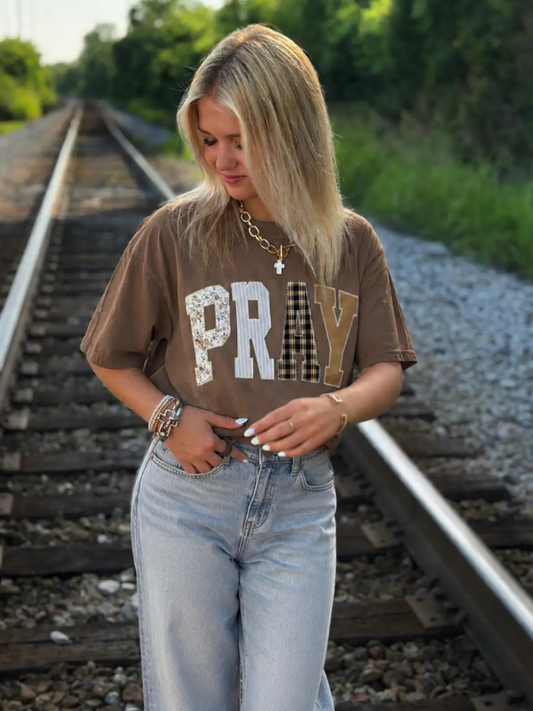 A woman stands on railroad tracks wearing a brown graphic t-shirt with the word 'PRAY' in textured lettering, paired with light-wash jeans.
