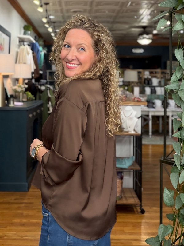 A woman with curly blonde hair wearing a brown satin blouse and blue jeans, looking back over her shoulder in a retail store setting.