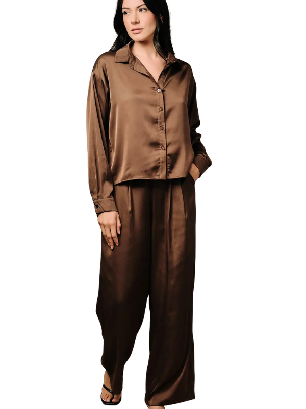 A woman modeling a matching brown satin pajama set with a long-sleeve button-up shirt and wide-leg pants against a plain white background.