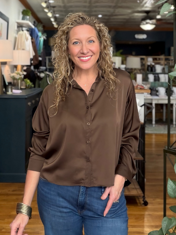 A smiling woman with curly blonde hair stands in a boutique wearing a brown satin button-down blouse and blue jeans.