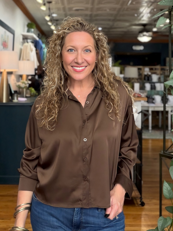 A smiling woman with curly blonde hair wearing a brown satin button-down shirt and blue jeans, standing in a retail store interior.