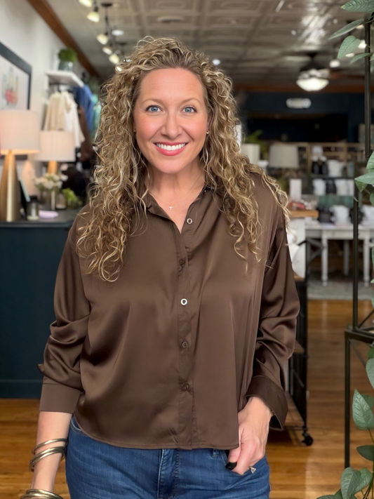 A smiling woman with curly blonde hair wearing a brown satin button-down shirt and blue jeans, standing in a retail store interior.