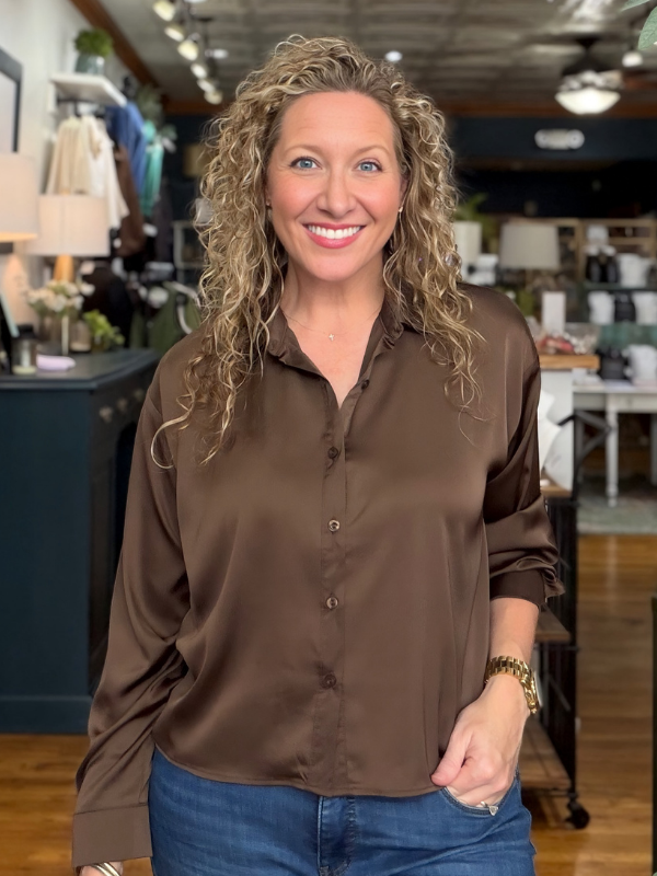 A smiling woman with curly blonde hair wearing a brown button-down satin blouse and blue jeans, standing inside a retail store.