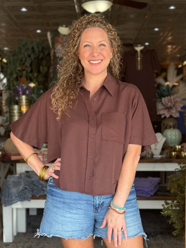 Woman wearing a brown shirt and denim shorts in an indoor setting with various items in the background.