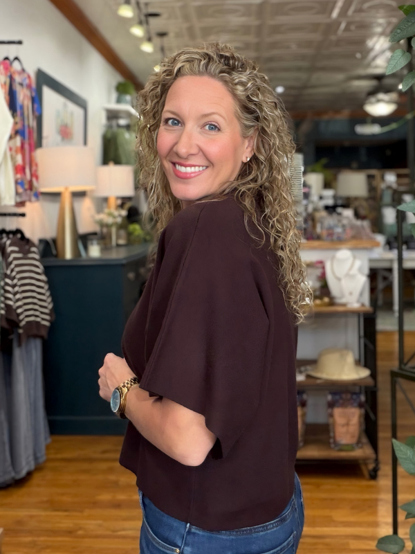 A smiling woman with curly blonde hair wearing a dark brown short-sleeve top and blue jeans, standing inside a retail store.