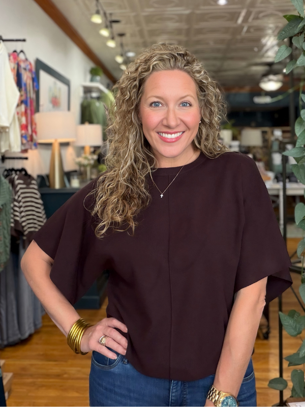 A smiling woman with curly blonde hair wearing a dark plum short-sleeve top and blue jeans, standing inside a boutique.