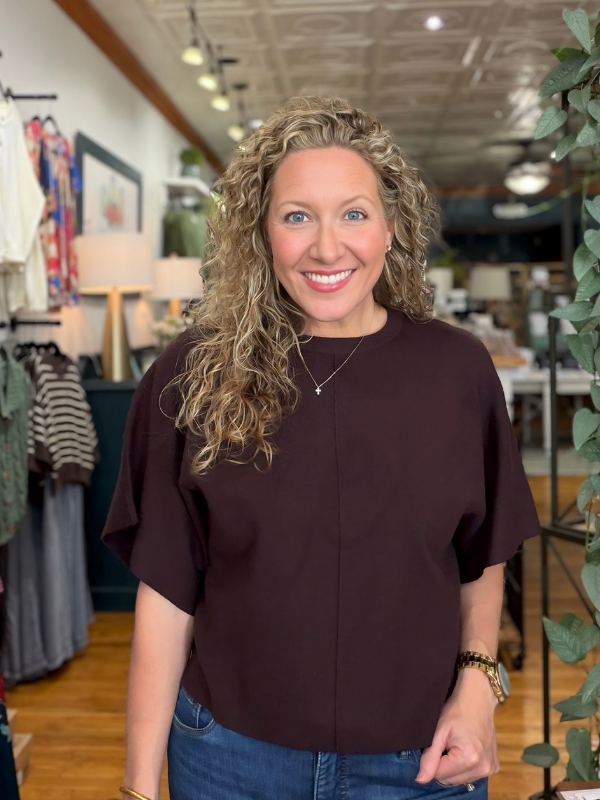 A smiling woman with curly blonde hair wearing a dark brown short-sleeve top and blue jeans, standing inside a retail store.