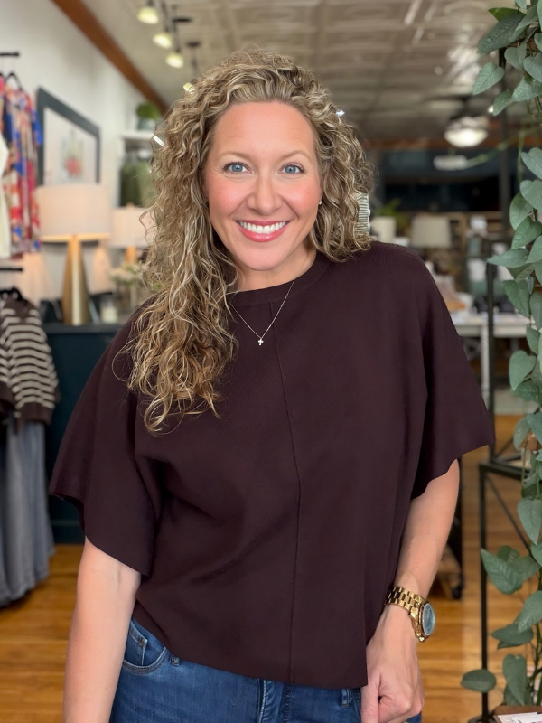 A smiling woman with curly blonde hair wearing a dark brown short-sleeve top and blue jeans, standing in a retail store interior.