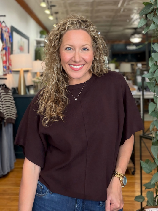 A smiling woman with curly blonde hair wearing a dark brown short-sleeve top and blue jeans, standing in a retail store interior.