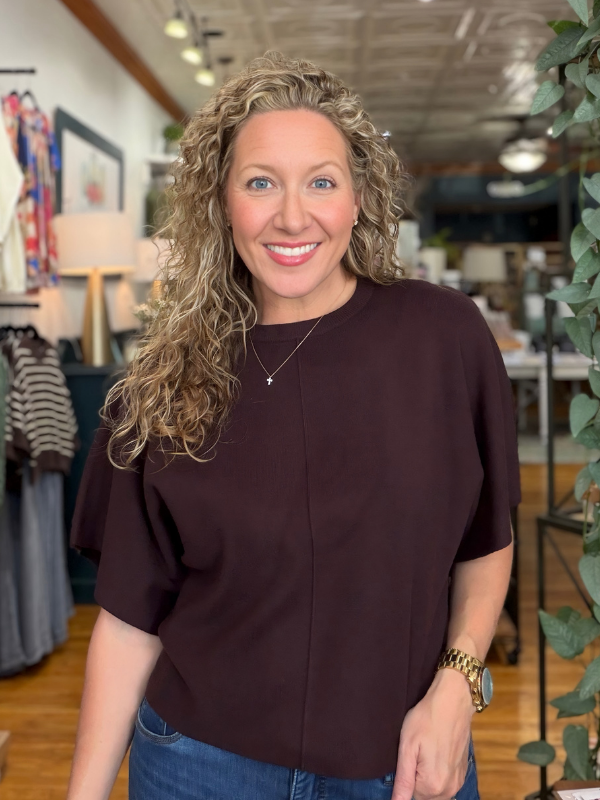 A smiling woman with curly blonde hair wearing a dark plum short-sleeve top and blue jeans, standing inside a boutique store.