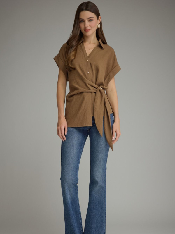 A woman modeling a brown short-sleeve button-front shirt with a self-tie waist, paired with blue jeans, against a solid gray studio background.