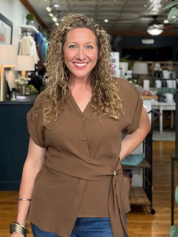 A smiling woman with curly blonde hair wearing a brown short-sleeve wrap-style blouse with a tie waist, standing in a retail store interior.