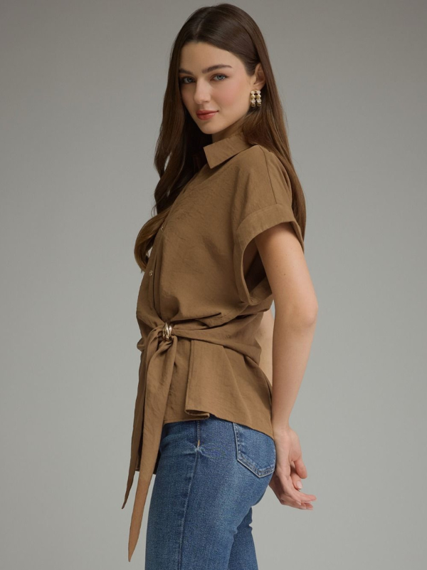 A woman modeling a brown short-sleeve collared blouse with a self-tie waist belt, paired with blue jeans, against a solid gray studio background.
