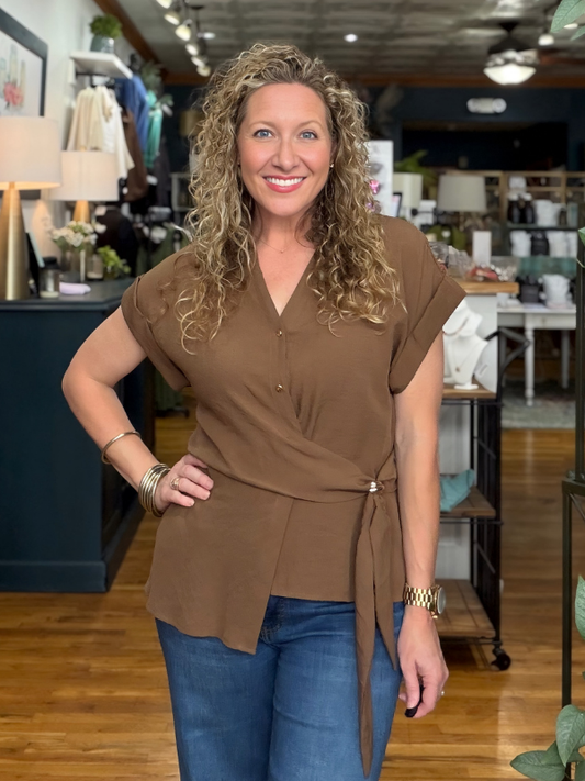 A woman with curly blonde hair wearing a brown short-sleeve wrap-style top with a tie waist and blue jeans, standing in a retail store interior.