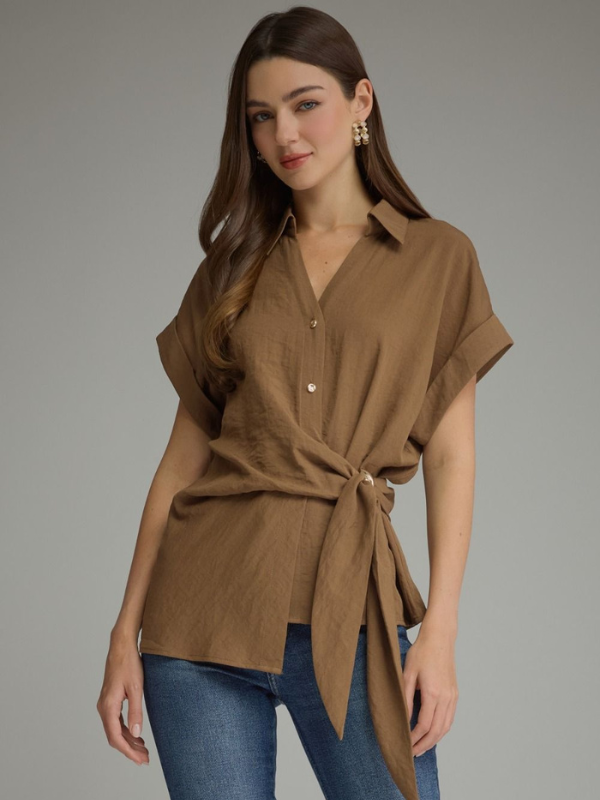 A woman modeling a brown short-sleeve button-up shirt with a tie waist, paired with blue jeans, against a solid gray studio background.
