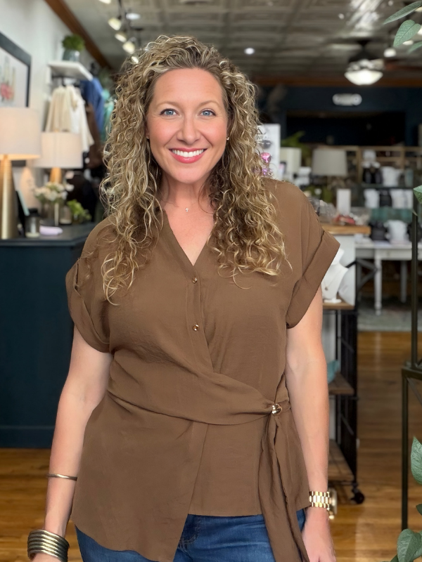 A smiling woman with curly blonde hair wearing a brown short-sleeve wrap-style blouse with a tie waist, paired with blue jeans, standing inside a retail store.