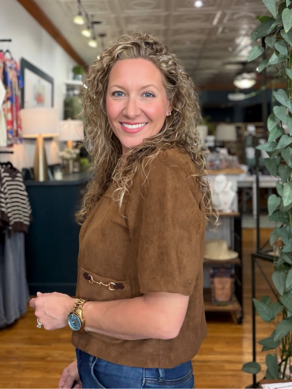 A smiling woman with curly blonde hair wearing a brown short-sleeve suede-like top and blue jeans, standing in a retail store interior with shelves, plants, and decor visible in the background.