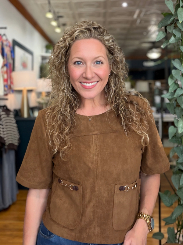A smiling woman with curly blonde hair wearing a brown suede short-sleeve top with front pockets, standing in a retail store interior.